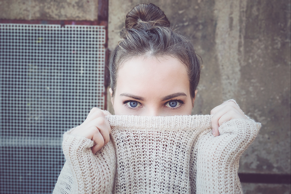 Industrial looking concrete and metal grid background. In front of it a close-up of a white person wearing an off-white knit sweater. They are pulling the sweater up with both hands so that half their face is hidden behind it. Their bright blue eyes are still visible.