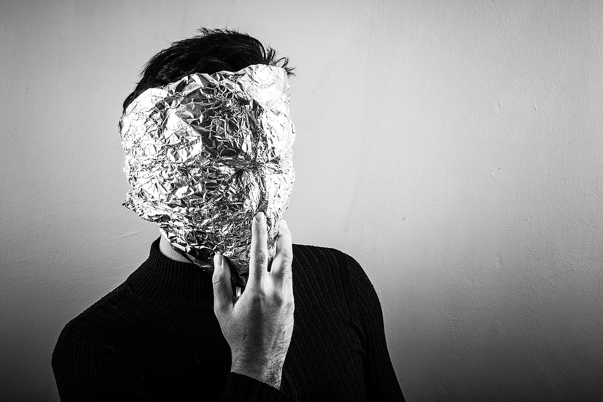 Black and white image of a person with short black hair, wearing a black sweater. Their face is covered by a mask made from crumbled up aluminum foil with no facial features. They are touching their chin.