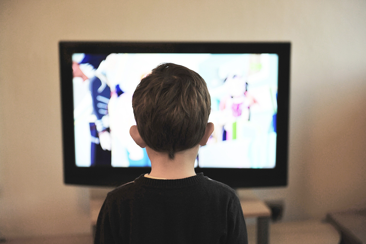 Young white child with short hair, wearing a black sweater, facing away from the camera, standing in front of a TV.