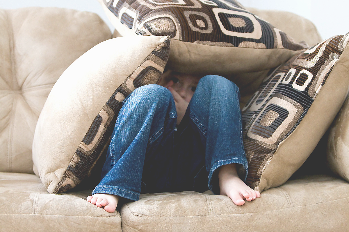 Beige sofa with beige and brown pillows in the shape of a cave. In the pillow cave a young white child is hiding. They are barefoot.