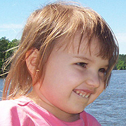 Image depicts Katie McCarron - a young Caucasian girl with medium long brown hair, squinting against the sun and biting her lower lip, holding back a cheeky smile.