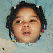 Image depicts Shylea Myza Thomas - a young African American girl with her black hair in many tiny braids, laying in bed holding her stuffed teddy bear.