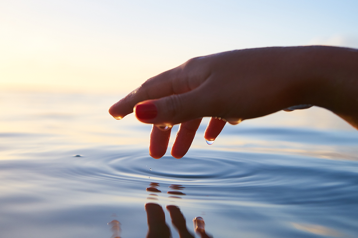 A hand reaching out over still water, drops hanging from its thumb. There are some circular ripples on the surface of the water where a finger was dipped into it.