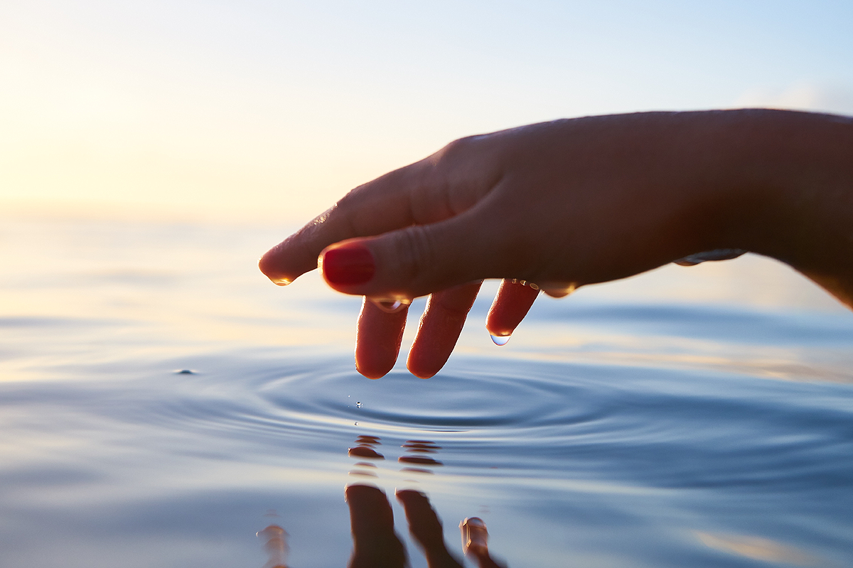 Eine Hand, die sich über stilles Wasser ausstreckt. Einzelne Wassertropfen hängen von ihrem Daumen. Dort, wo die Finger das Wasser berührt haben, ziehen sich kreisförmige Wellen über die Wasseroberfläche.