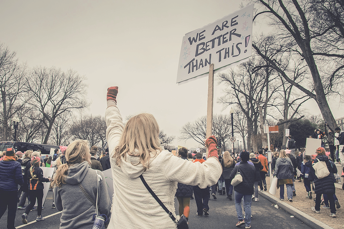 A group of people on the streets, marching, protesting. A white person with their back to the camera wearing a white jacket has their left fist raised into the air. In their right hand, they carry a sign saying "We are better than this!"