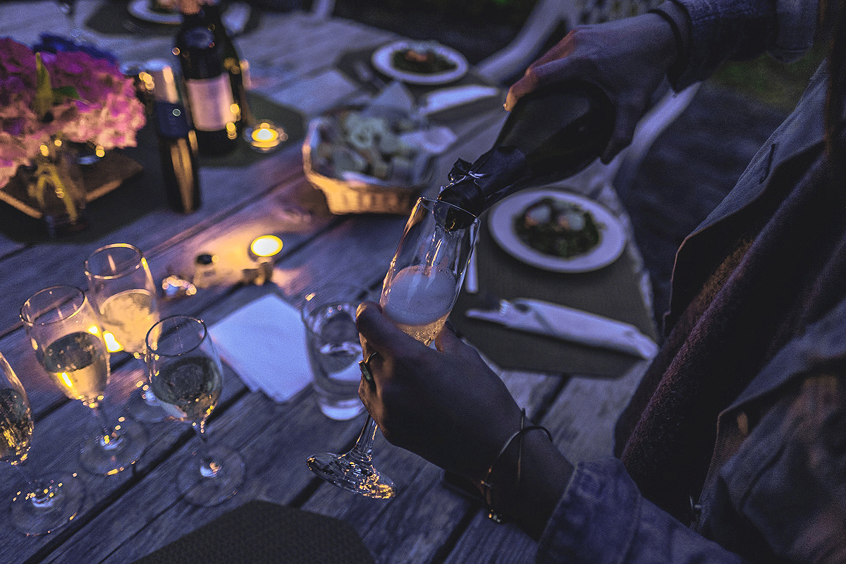 A pair of hands, one holding a glas, the other pouring champagne into it. In the blurry background a table, fully set, ready to sit down and eat at. A lit candle and dark colors suggest it's a night scene.