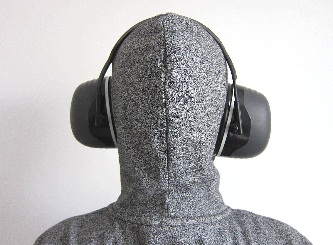 Autistictic from behind, wearing a grey hoodie with the hood up. They are wearing the Peltor X5A ear defenders showing how thick the shells are and how far they stick out which is a lot.