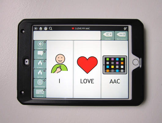 Ein AAC Gerät in einer schwarzen Hülle. Der Bildschirm zeigt drei große Buttons: I, LOVE, AAC - mit den dazugehörigen Symbolen.