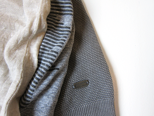 Close-up of three pieces of clothing. The first is beige linen, the second is grey cotton with black pinted on stripes, the third is grey cotton with a square knit texture.