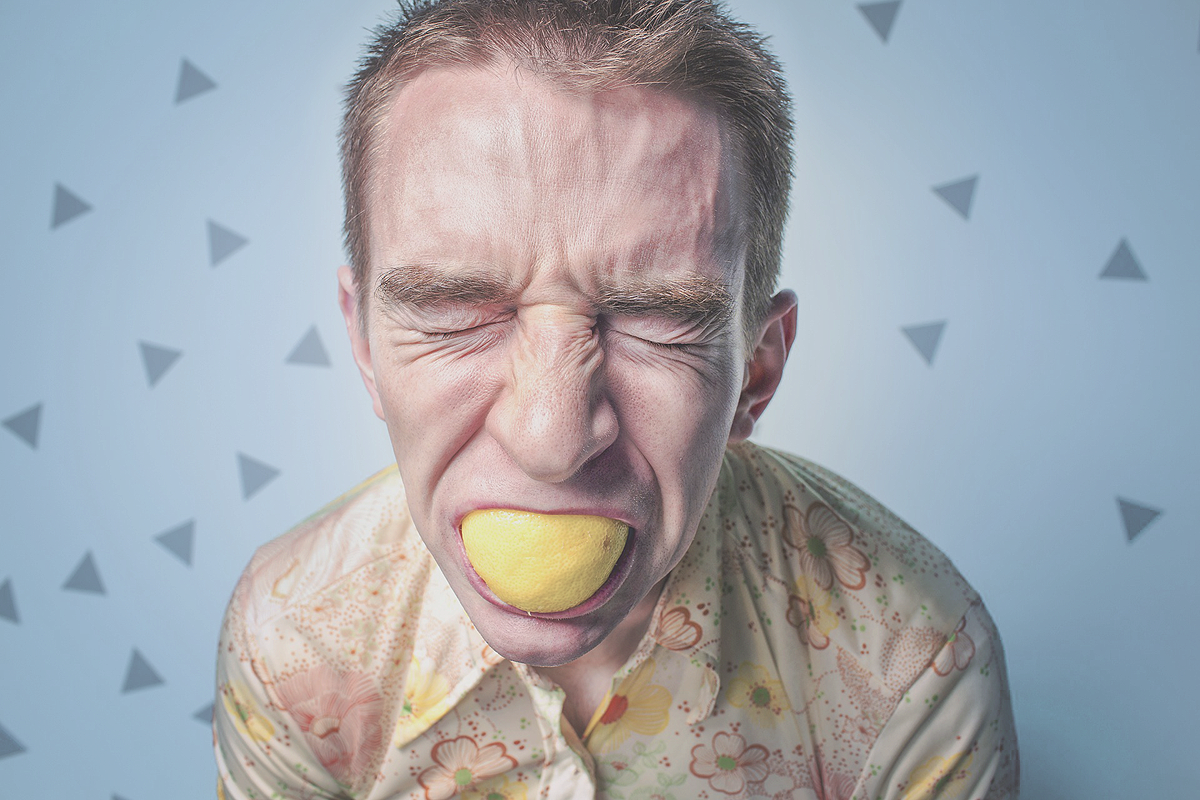 A person with short blonde hair biting into a lemon. Their face is all scrunged up, showing how sour that piece of food is.