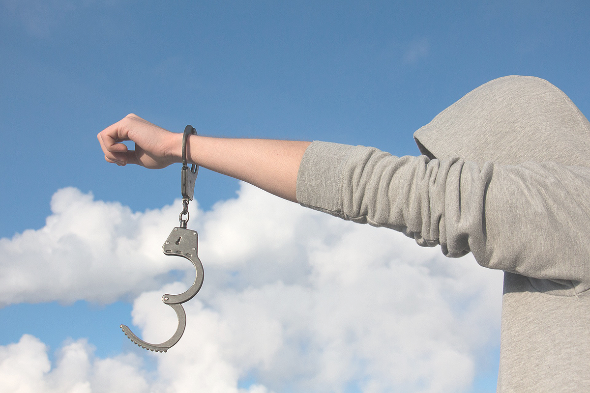 Blue sky with some whity clouds in the background. A white person wearing a grey hoodie with the hood hiding their face in the front. They have their fist raised with handcuffs broken open dangling from their wrist.