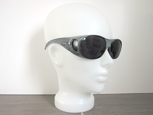 White background, grey floor, white glass head wearing large grey sunglasses with dark grey lenses.