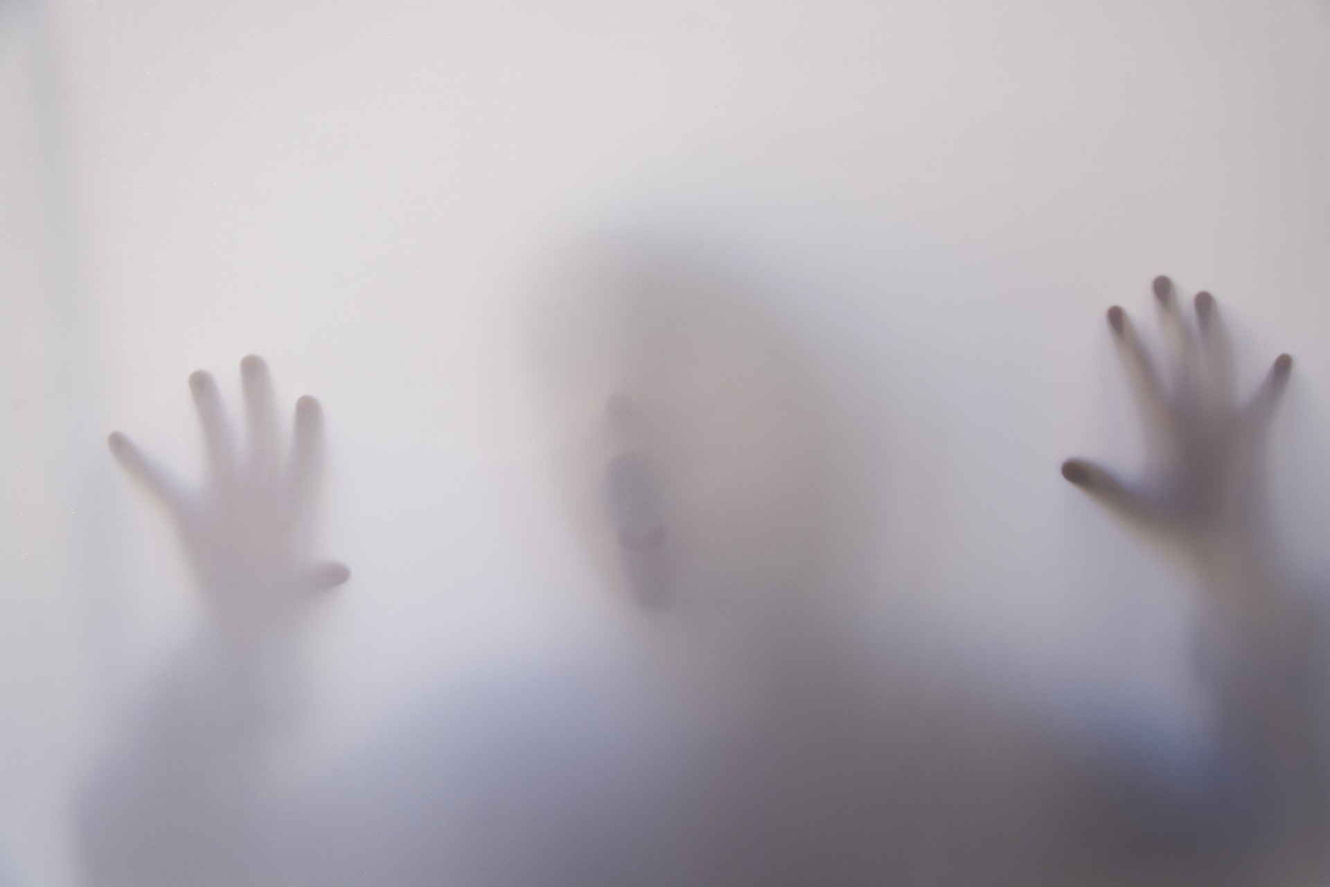 Milky glass with the blurry figure of a child behind it. Both hands are resting against the glass, their face is close to it as well, their mouth is open as if struggling for air.