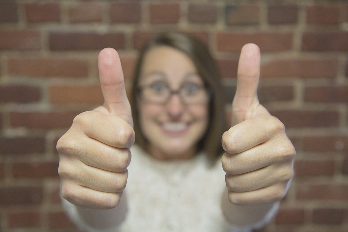 A close-up of two hands in fists giving a double thumbs-up. In the background a blurry person with chin-long hair, wearing glasses, standing in front of a brick wall.