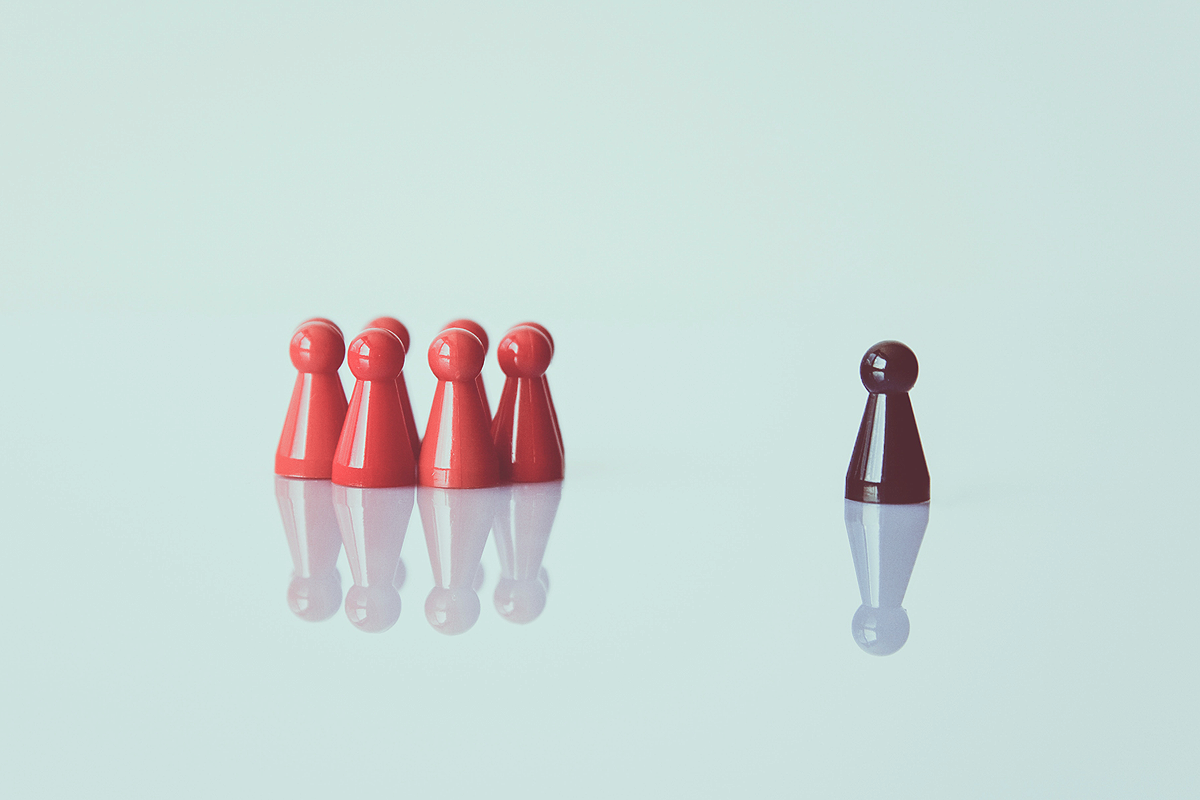 Blueish grey background with a group of red game pieces standing together in a group. On their right, a bit away, stands a lonely black game piece.