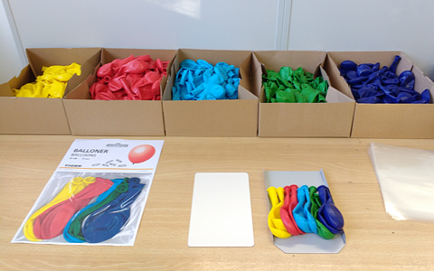 A desk with 5 boxes on it. Each box is filled with a different color of balloon: yellow, red, light blue, green, dark blue. In the front there's a paper with a photograph showing the order of the colors the balloons should be packaged. Right next to it a board with a row of balloons assembled in the correct color order, ready to be inserted into their packaging.