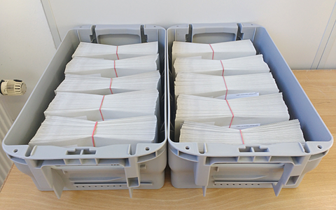 A desk with two grey post boxes on it. Both boxes are filled with hundreds of envelopes bundles into packets with red rubber bands.