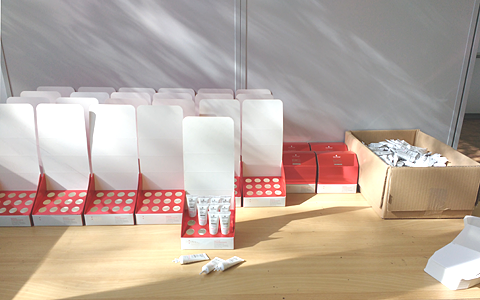 A desk filled with rows of folded packaging with spaces for tubes. On the right a box filled with tubes. One package is being filled with the tubes, some are already placed into the matching spaces. A couple of tubes are laying on the desk.