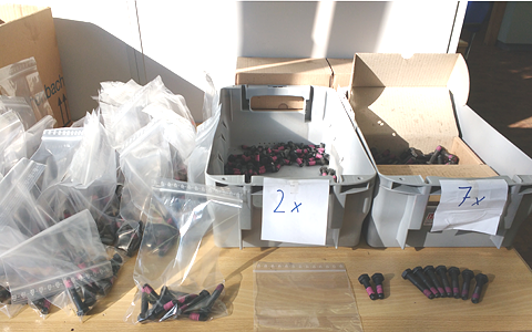 A desk with two grey boxed full of two sizes of screws on it. Left of the boxes are dozens of readily packaged plastic bags filled with screws. In the front an example of a filled bag, an empty plastic bag, two small, and seven large screws.