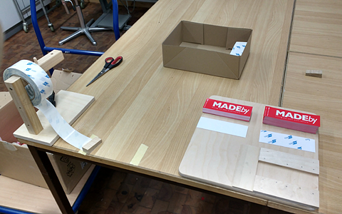 A desk with a box of pre-printed stickers. On the right side there's a contraption attached to the desk that helps a person tear pieces of adhesive tape off a large roll. On the desk there is a wooden board that helps a person attach the adhesive side to the stickers.