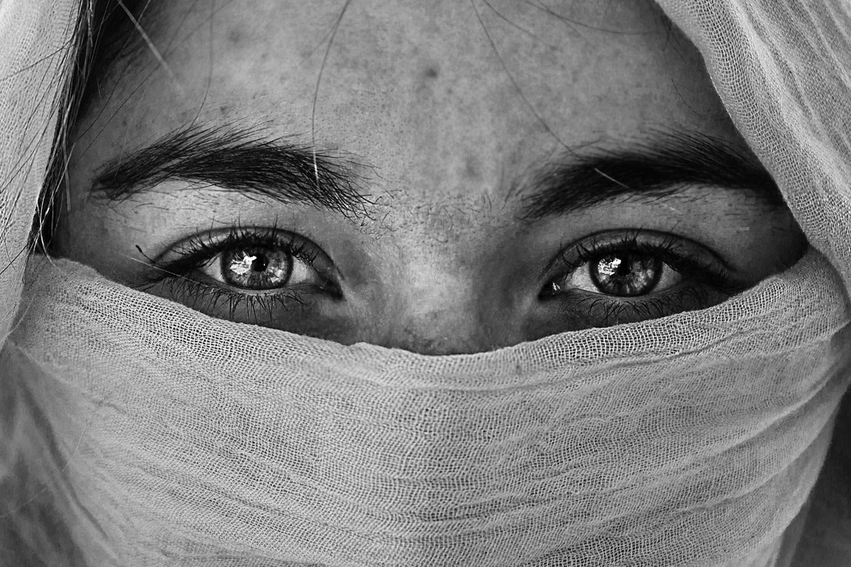 Black and white photograph of a pair of eyes, with thick, bushy, black eyebrows, centered in the frame, staring directly into the camera. The rest of the face is hidden behind a head scarf.