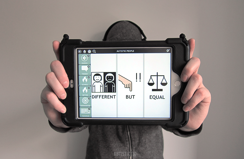 Autistictic holding their black AAC tablet into the camera. The three large buttons on the screen read "DIFFERENT", "BUT", "EQUAL". Autistictic is wearing a grey hoodie. The AAC tablet is hiding their face.
