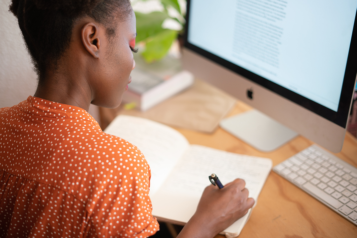 Dark-skinned person wearing an orange patterned shirt sitting at a desk. They have a computer monitor in front of them with a site open and are taking notes in a notebook.