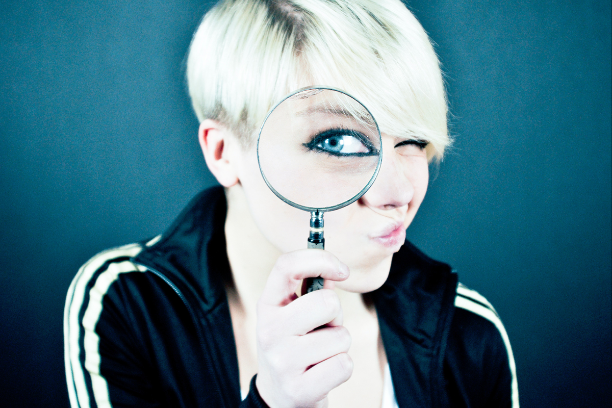Person with short blonde hair with a magnifying glass infront of their eye. The eye is magnified while the rest of their face is normal sized.