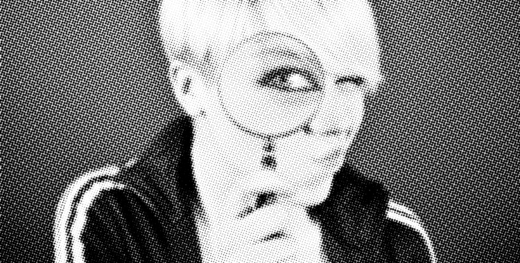 Person with short blonde hair with a magnifying glass infront of their eye. The eye is magnified while the rest of their face is normal sized.