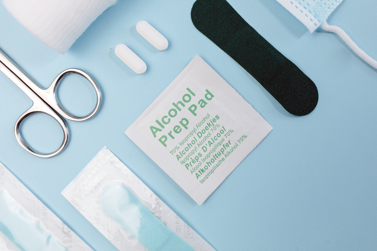 A light blue surface with several different first aid items on it: an alcohol pad, a bandaid, some pills, and some scissors.